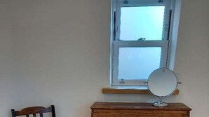 A wooden dresser with drawers and a mirror, a wooden chair with a cushion, and a window above the dresser. - Holiday Cottage Sleeps 3 With Garden (Prestwick)