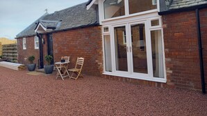 A brick house with a large window, a door, and a small patio with chairs and planters. - Holiday Cottage Sleeps 3 With Garden (Prestwick)