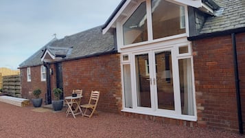 A brick house with a large window, a door, and a small patio with chairs and planters.