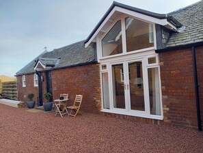A brick house with a large window, a door, and a small patio with chairs and planters. - Holiday Cottage Sleeps 3 With Garden (Prestwick)