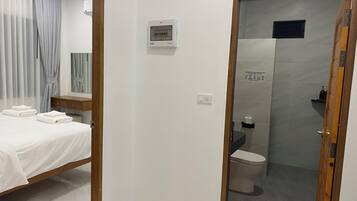 A hotel room with a bed, a bedside table, a mirror, a wall-mounted air conditioning unit, and a shower area with a toilet.