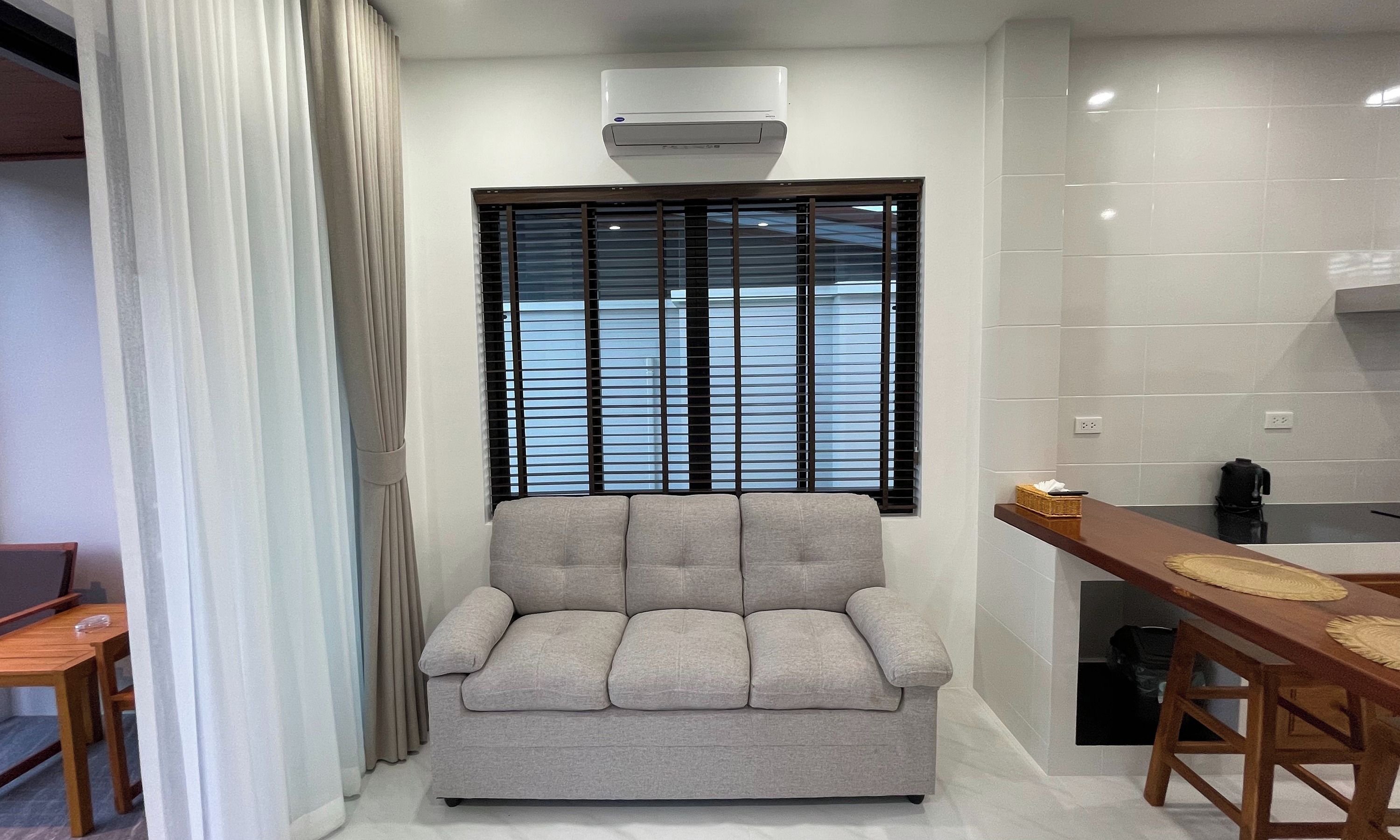 A modern living room with a grey sofa, a window with blinds, a ceiling-mounted air conditioner, and a wooden dining table with a basket.