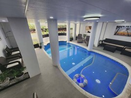 An indoor swimming pool with a blue interior, surrounded by white pillars, and a seating area with chairs and a bench.