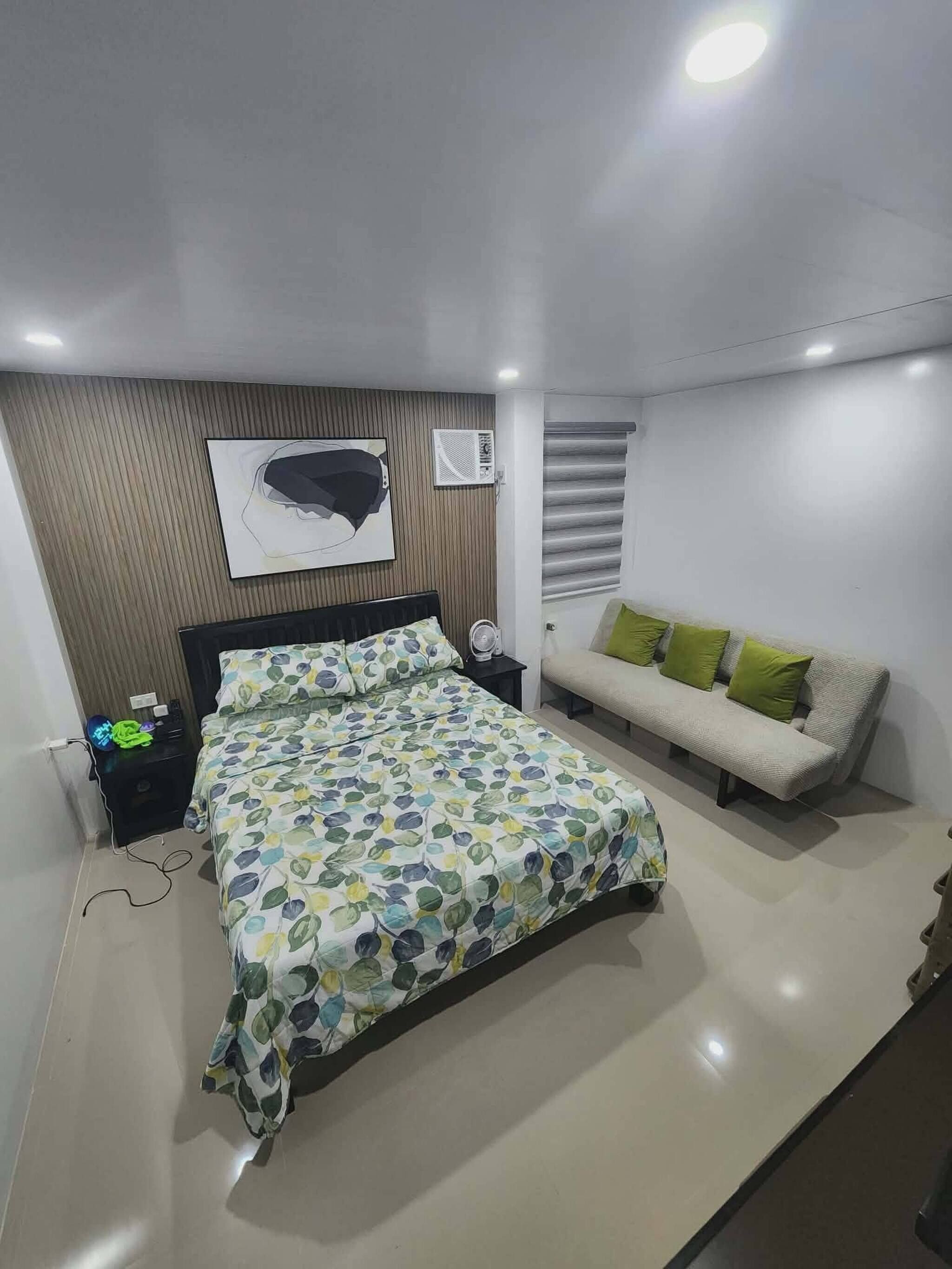 A modern bedroom with a bed, a nightstand, a sofa, and a wall-mounted air conditioner.