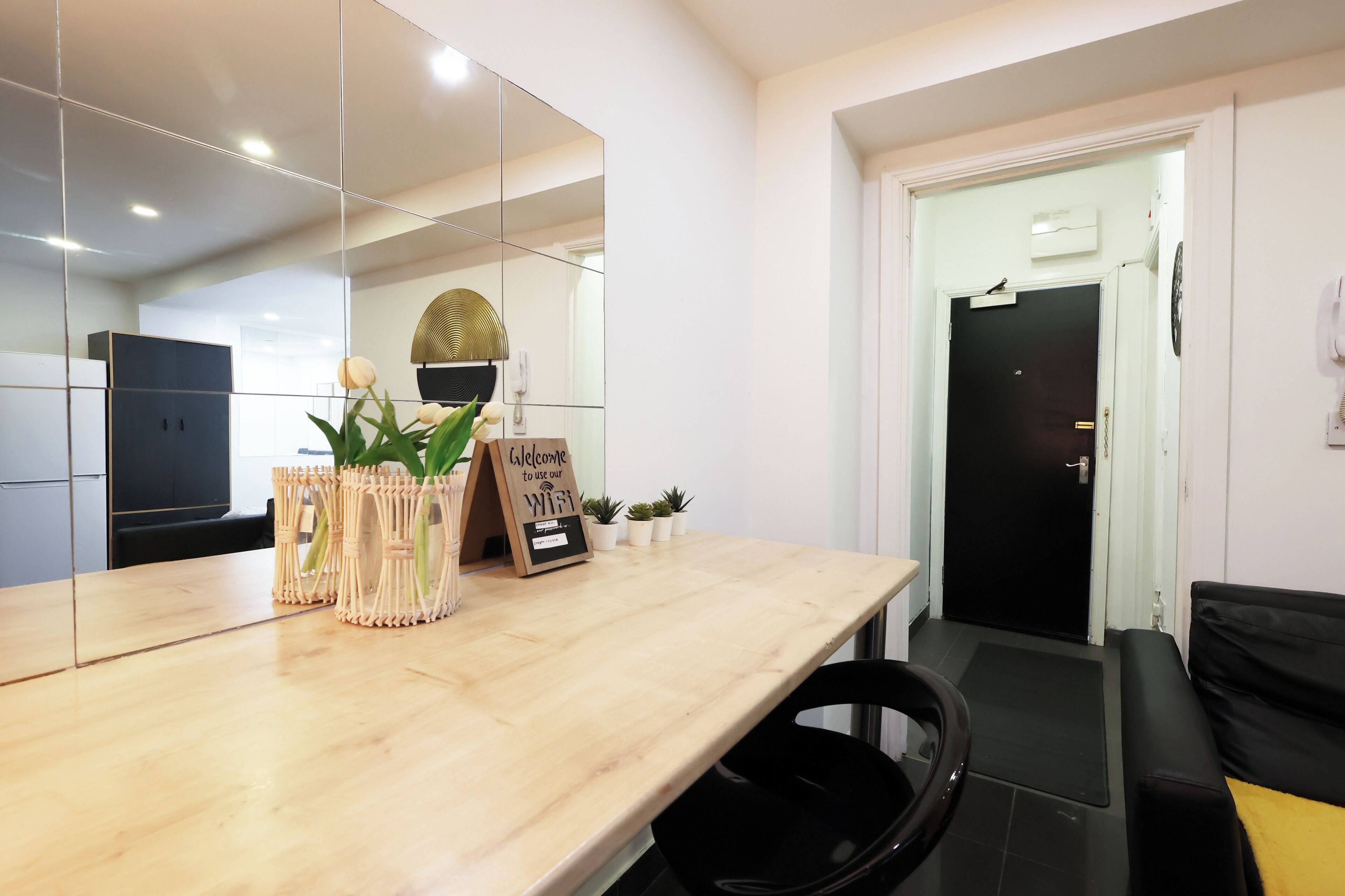 A modern hotel room with a wooden reception desk, a black chair, a black sofa, a small potted plant, a welcome sign, and a door with a black handle.