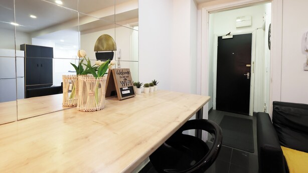 A modern hotel room with a wooden reception desk, a black chair, a black sofa, a small potted plant, a welcome sign, and a door with a black handle. - Walk to Stadium and City Center (Cardiff)