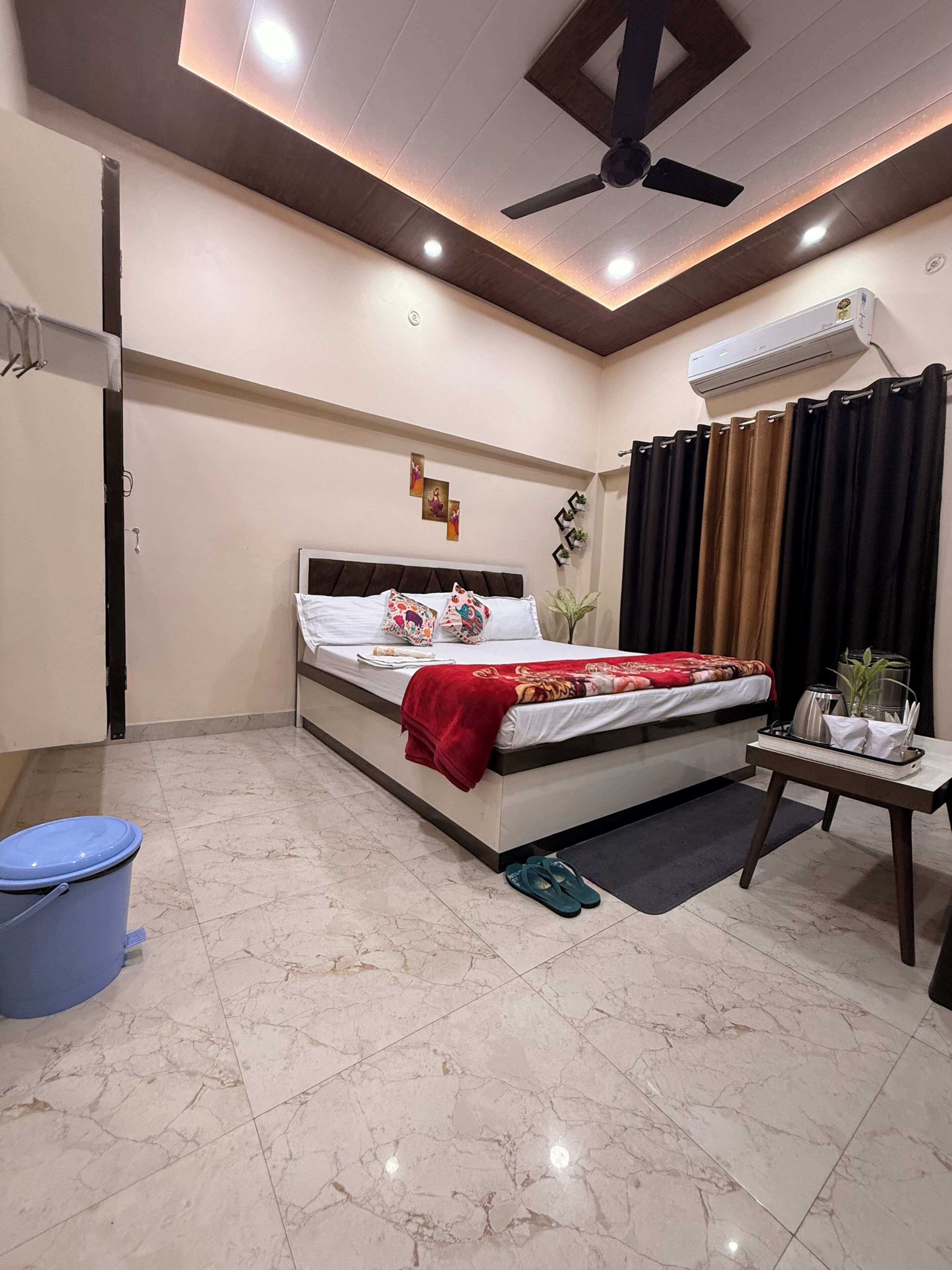 A hotel room with a bed, a ceiling fan, an air conditioning unit, a small table with a tray, and a blue trash bin.