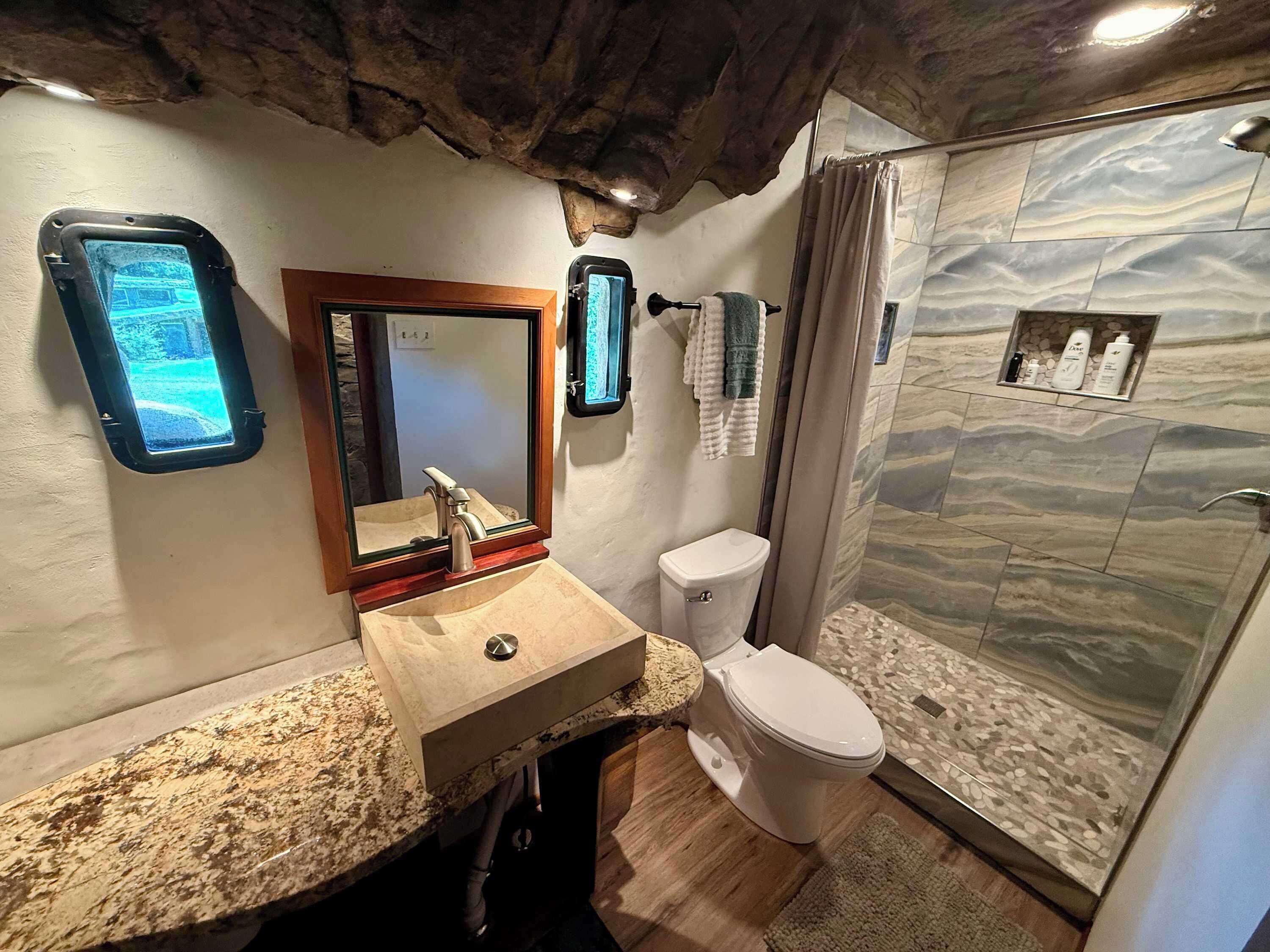 Une salle de bain avec un lavabo surmonté d’un plan de travail en granit, un miroir, des toilettes et une douche dont le mur est recouvert de carreaux imitant la pierre.