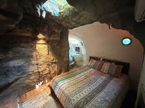 Un dormitorio con paredes de piedra, una cama, un televisor pequeño y un ventanal circular.