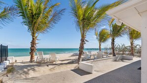 Un patio frente a la playa con sillas blancas, palmeras y vistas al océano.