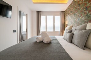 A hotel room with a bed, a flat-screen TV mounted on the wall, a window with curtains, and a patterned wallpaper on the wall. - Sol Atelier | Apartments by Monaro* * * * * (Madrid)
