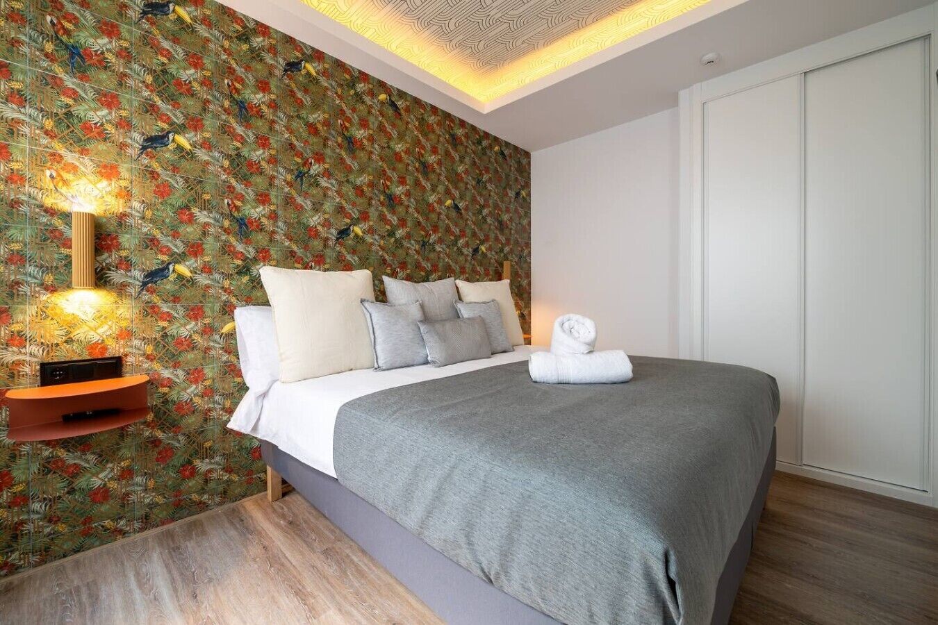 A bedroom with a bed, pillows, a bedside table with a lamp, and a wall with a floral pattern.