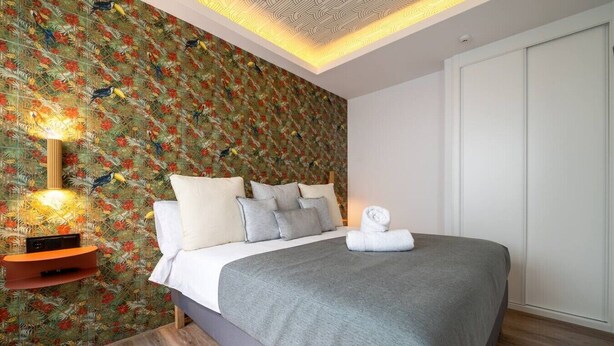 Et soveværelse med seng, puder, et natbord med lampe, og en væg med blomstermønster.