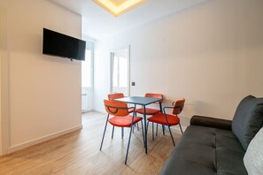 A modern living room with a flat-screen TV mounted on the wall, a dark sofa, and a small dining table with red chairs. - Sol Atelier | Apartments by Monaro* * * * * (Madrid)
