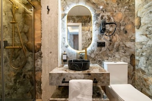 Ein Badezimmer mit Marmorwand, einem spiegel mit goldener Rahmung, einem schwarzen Waschbecken, einer weißen Toilette und einer Dusche mit goldfarbener Brauseköpfe.