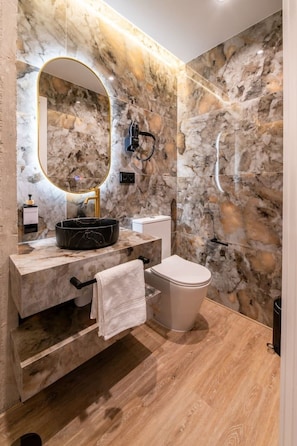 A modern bathroom with a marble wall, a round mirror, a black sink, a toilet, and a towel rack. - Sol Atelier | Apartments by Monaro* * * * * (Madrid)