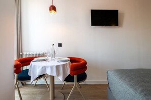 A hotel room with a dining table set for two, a flat-screen TV mounted on the wall, and a bed with a grey cover. - Sol Atelier | Apartments by Monaro* * * * * (Madrid)