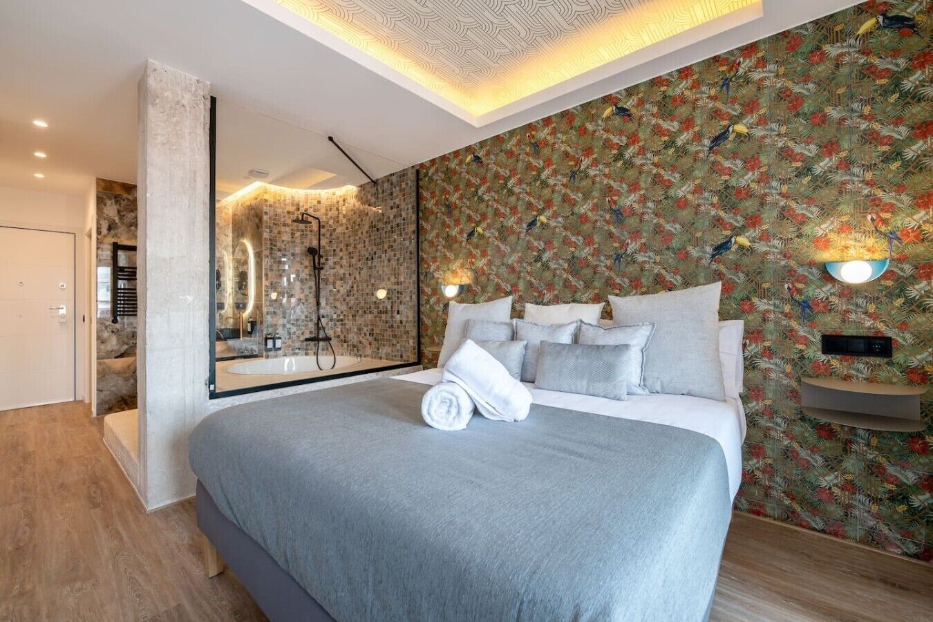 Un dormitorio moderno con una cama grande, un baño con ducha de vidrio y una pared con motivos florales tropicales.