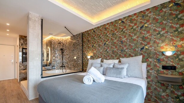A modern bedroom with a large bed, a bathroom with a glass-enclosed shower, and a wall with a tropical floral pattern. - Sol Atelier | Apartments by Monaro* * * * * (Madrid)