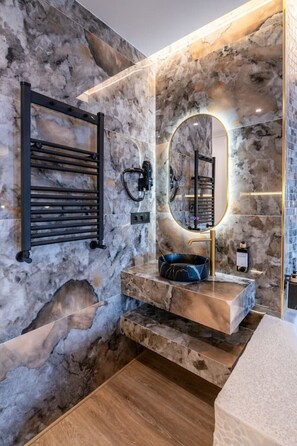 A bathroom with a marble wall, a black towel rack, a round mirror with a gold frame, and a wooden floor. - Sol Atelier | Apartments by Monaro* * * * * (Madrid)
