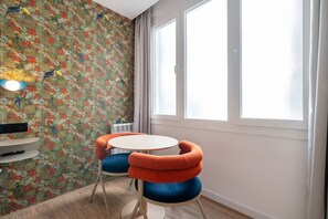 Ein Zimmer mit tropischem Wandpapier, einem runden Tisch und zwei Stühlen mit orange- und blauen Kissen.