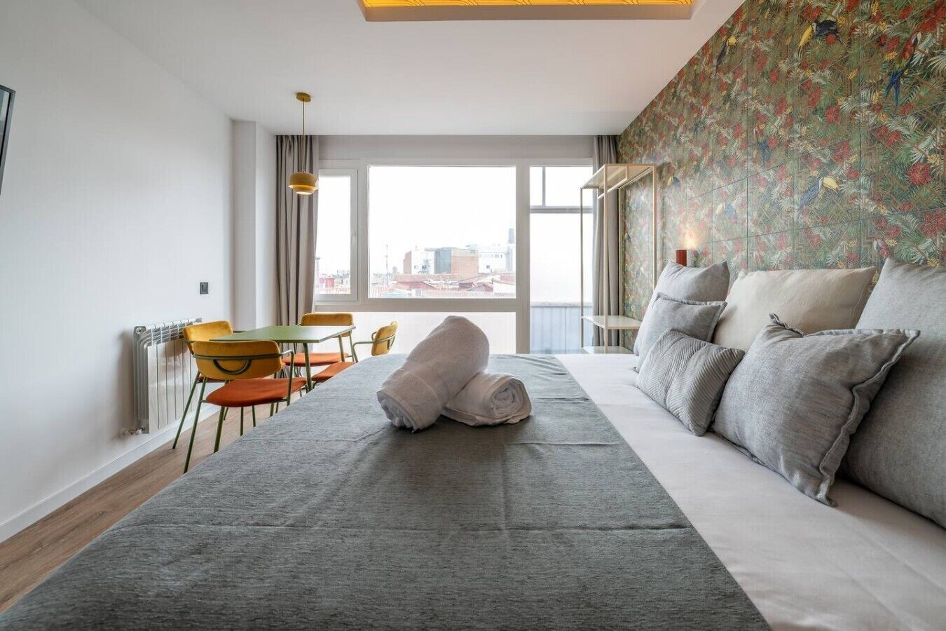 Una habitación de hotel con una cama, una mesa de comedor con sillas, un radiador y un ventanal con vistas a la ciudad.