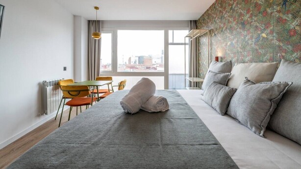 A hotel room with a bed, a dining table with chairs, a radiator, and a large window with a city view. - Sol Atelier | Apartments by Monaro* * * * * (Madrid)