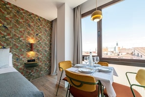 A hotel room with a bed, a dining table set for two, a city view through large windows, and a wall with a tropical wallpaper. - Sol Atelier | Apartments by Monaro* * * * * (Madrid)