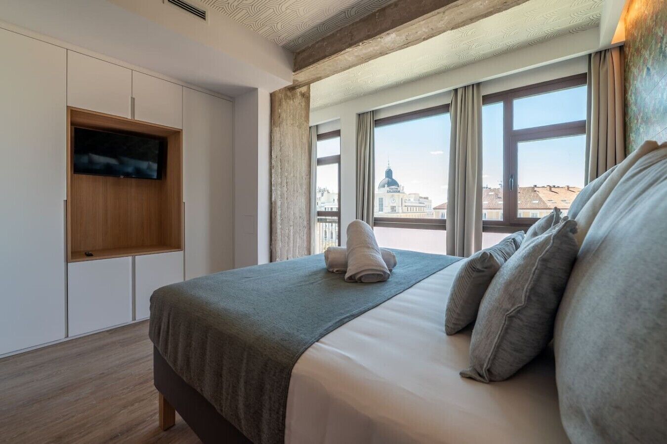 A hotel room with a bed, a flat-screen TV, and a view of buildings through large windows.