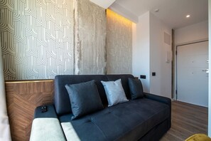 A modern living room with a dark sofa, patterned wallpaper, and a wooden accent wall. - Sol Atelier | Apartments by Monaro* * * * * (Madrid)