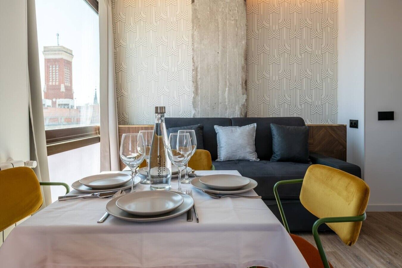 Un coin repas avec une table dressée pour quatre personnes, une bouteille d’eau et une vue sur un bâtiment par la fenêtre.