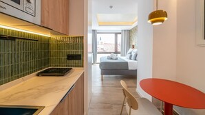 A modern kitchen with a microwave, a stove, and a green tiled backsplash. There is a dining area with a red table and a chair. A bed with a gray blanket and pillows is visible in the background. - Sol Atelier | Apartments by Monaro* * * * * (Madrid)