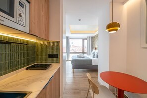 A modern kitchen with a microwave, a stove, and a green tiled backsplash. There is a dining area with a red table and a chair. A bed with a gray blanket and pillows is visible in the background. - Sol Atelier | Apartments by Monaro* * * * * (Madrid)