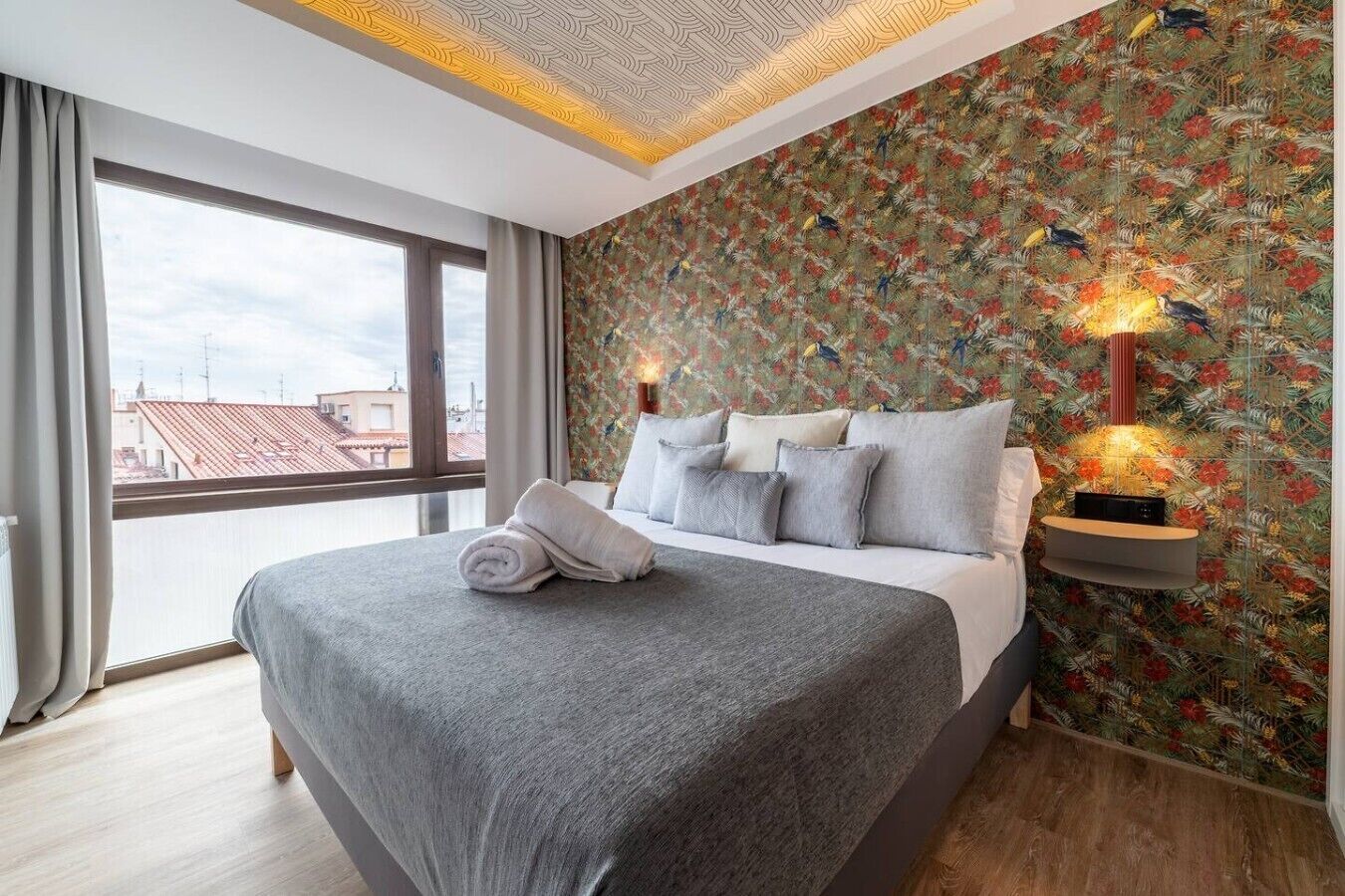 A hotel room with a large bed, a view through a window, and a patterned wall.