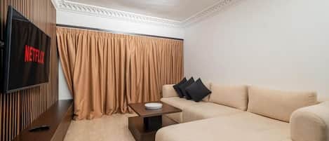 Una sala de estar moderna con un televisor de pantalla plana, un sofá color beige, una mesita de centro y una cortina.