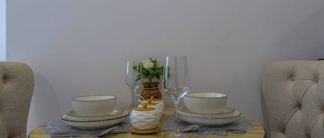 A round dining table with two white bowls and two wine glasses, a vase with a flower, and a small white pitcher on a wooden table with a lace doily.