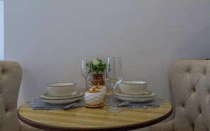 A round dining table with two white bowls and two wine glasses, a vase with a flower, and a small white pitcher on a wooden table with a lace doily. - Cozy Nest 1BR Kilimani (Nairobi)