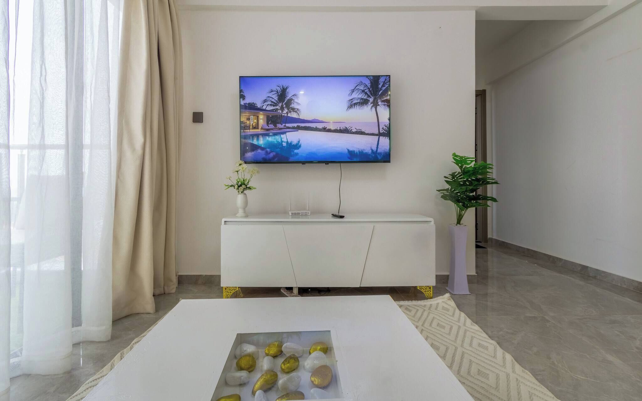 Una sala de estar moderna con un televisor de pantalla plana montado en la pared, un mueble blanco debajo, una mesa de centro con piedras decorativas y una planta en maceta al lado.