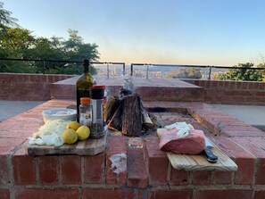 A brick outdoor cooking area with a fire, a bottle of olive oil, lemons, onions, and a meat block.