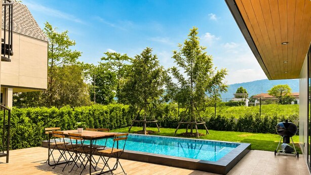 Un área de piscina moderna al aire libre con terraza de madera, una mesa de comedor con sillas y una parrilla.