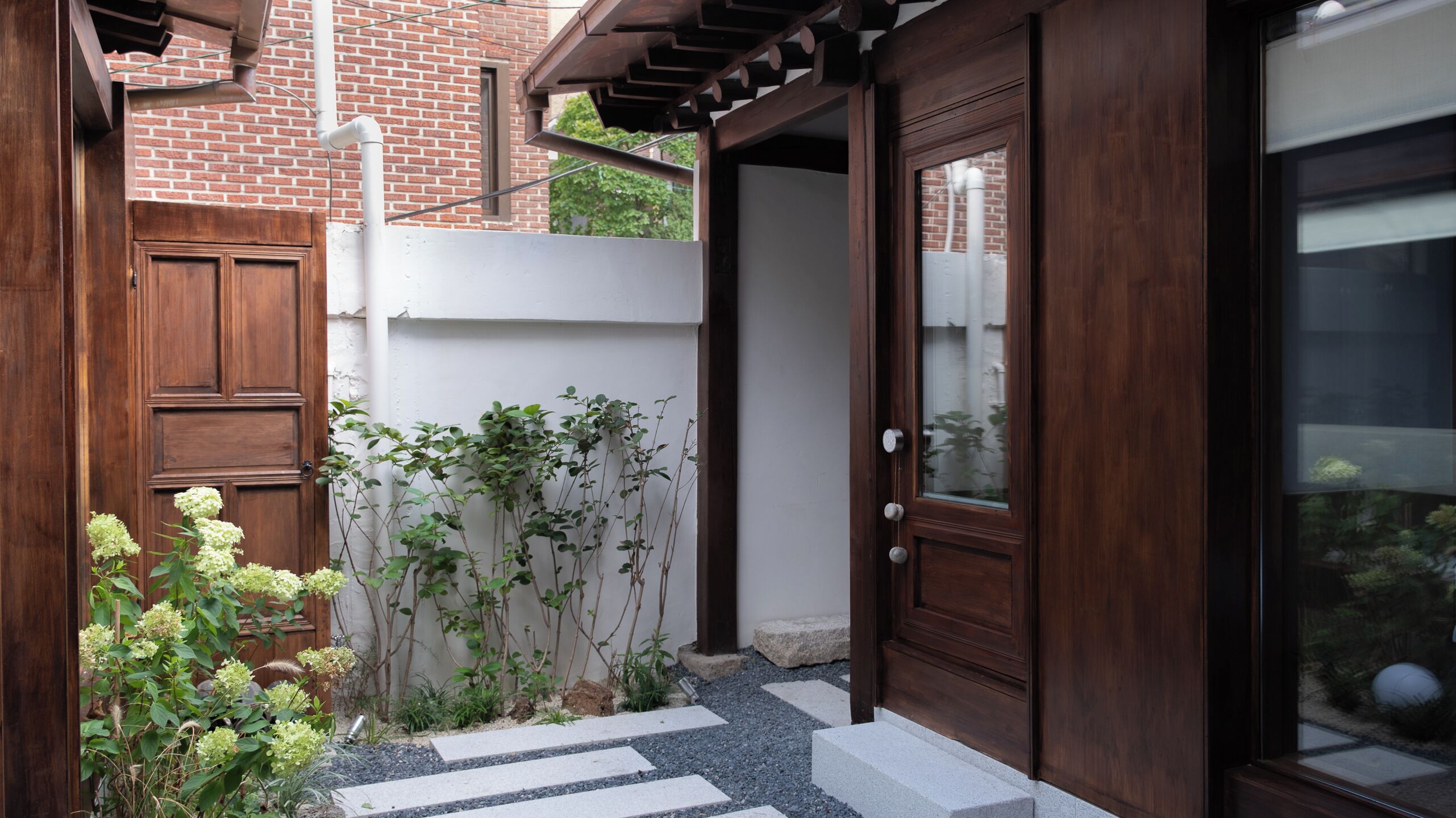 A wooden door with a glass panel, a brick wall, and a small garden with plants and a stepping stone path.