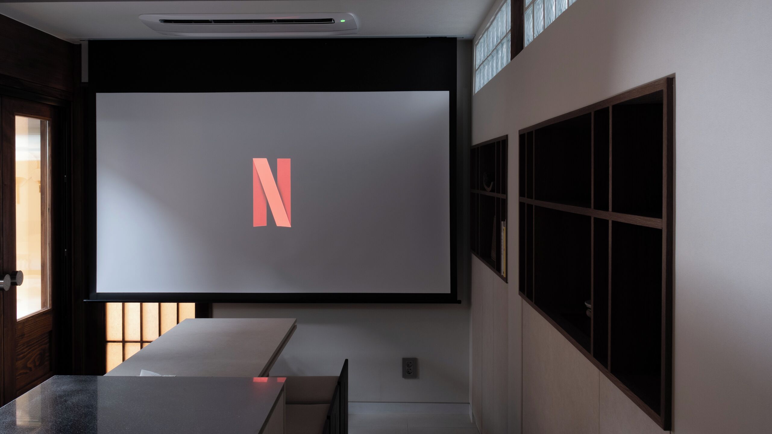 A modern interior with a large projection screen displaying a letter 'N', a minimalist table, and a window with blinds.