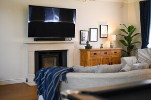 Ein Wohnzimmer mit Kamin, einem flachbildfernseher darüber, einer Couch, einer Holzkommode und gerahmten Bildern an der Wand.