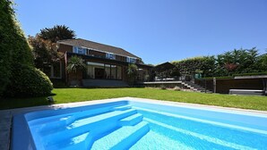 A swimming pool with a clear blue water surface, surrounded by a well-maintained lawn and a house with a front porch. - Milcourt -  a managed house that sleeps 8 guests  in 4 bedrooms (Canterbury)