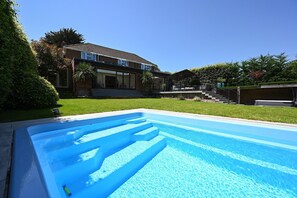 A swimming pool with a clear blue water surface, surrounded by a well-maintained lawn and a house with a front porch. - Milcourt -  a managed house that sleeps 8 guests  in 4 bedrooms (Canterbury)