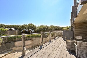 A wooden deck with a railing, overlooking a landscaped area with trees and a building with a closed shutter. - Pett Beach Landing -  a holiday let that sleeps 2 guests  in 1 bedroom (Hastings)