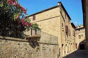 A narrow street with stone buildings, a balcony with a metal railing, and a clear blue sky. - A romantic house with a garden in Val d'Orcia. Between the past and the present. (Monticchiello)