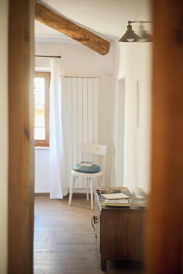 A room with a wooden floor, a white chair with a cushion, a small table with a book, and a radiator. - A romantic house with a garden in Val d'Orcia. Between the past and the present. (Monticchiello)