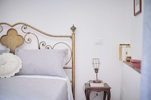 A neatly made bed with a metal headboard, a bedside table with a lamp, and a wall-mounted shelf with books. - A romantic house with a garden in Val d'Orcia. Between the past and the present. (Monticchiello)