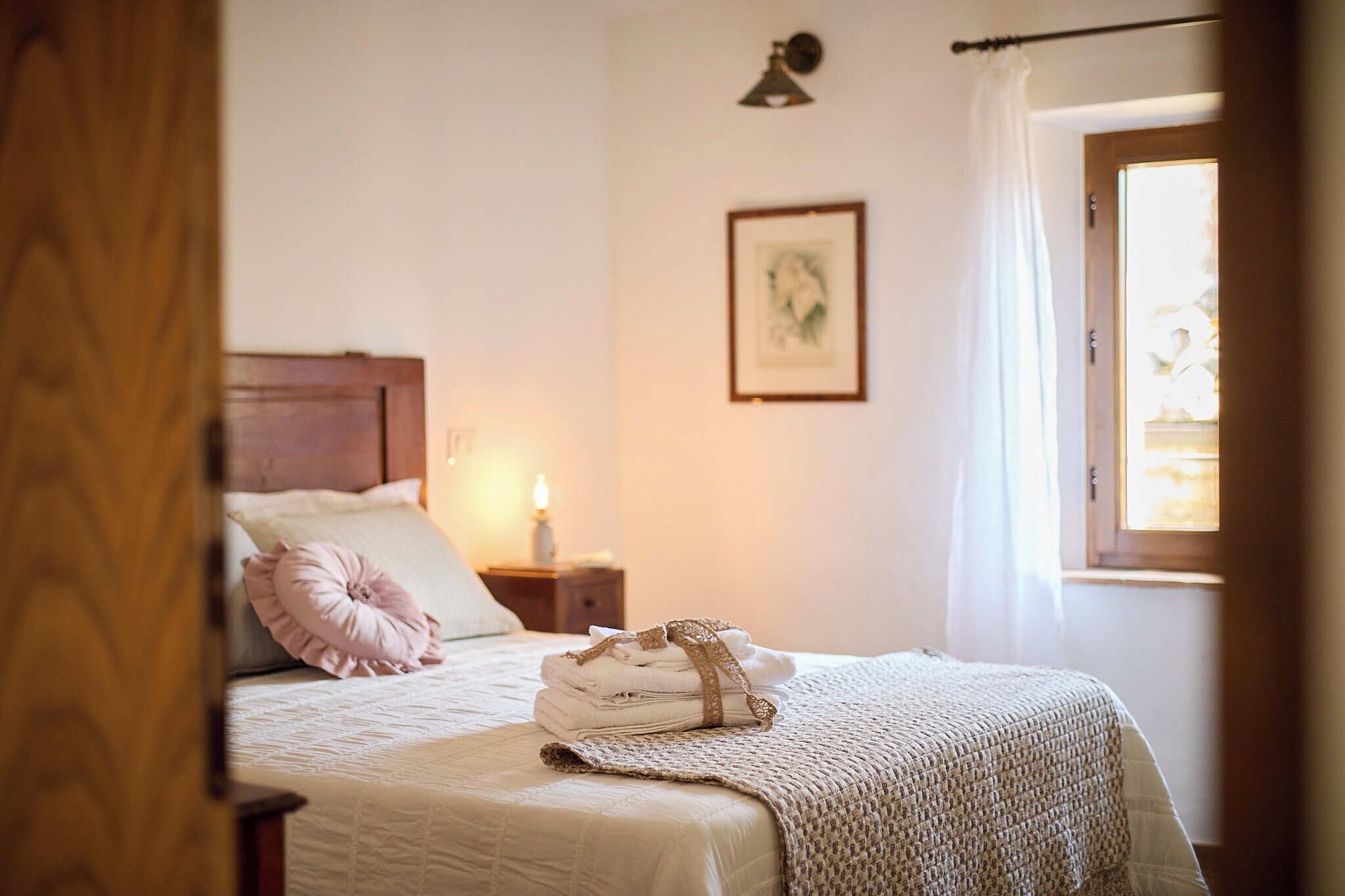 A bedroom with a bed, pillows, a bedside lamp, a framed picture on the wall, and a window with curtains.